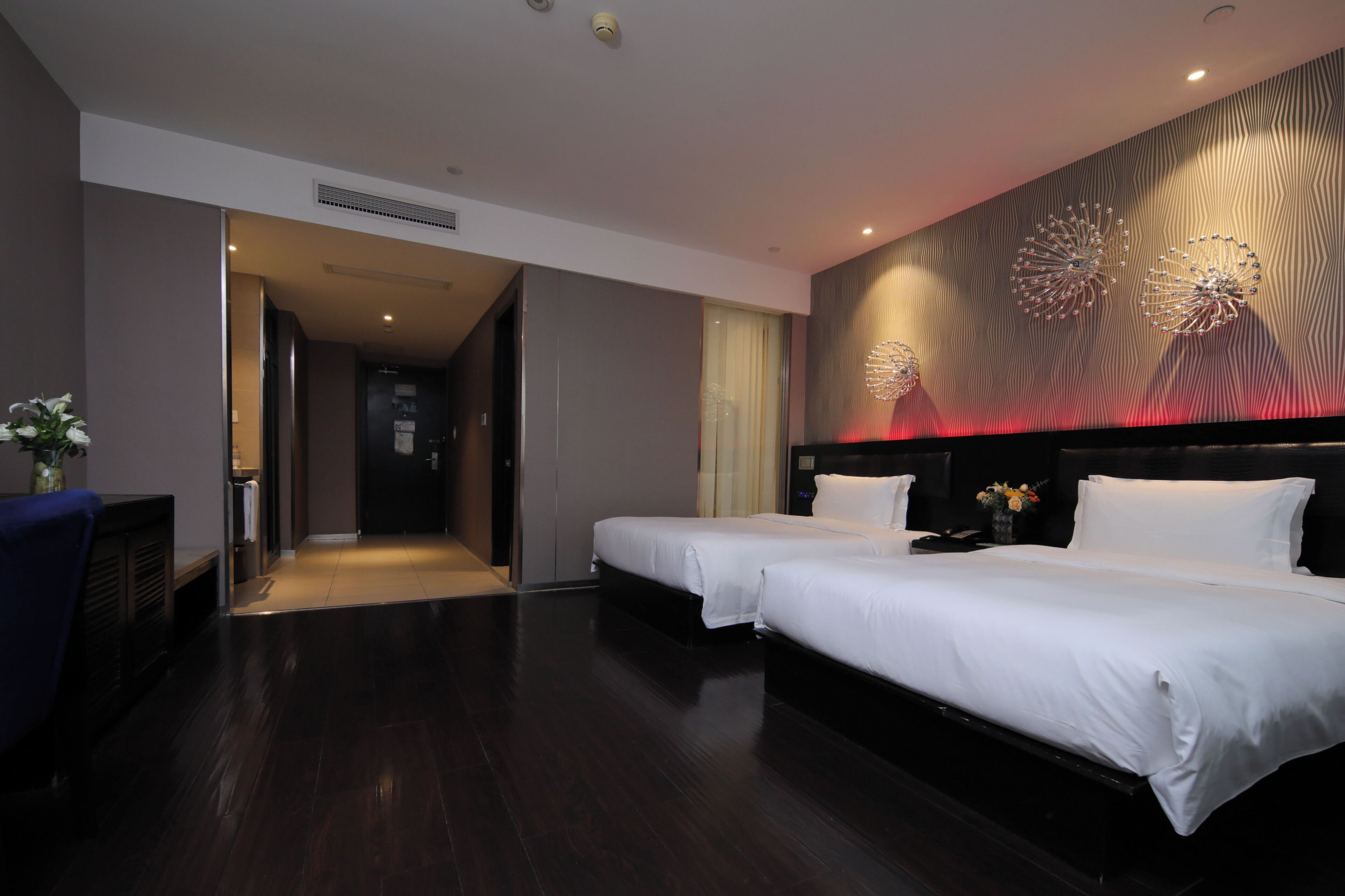 A hotel room with two beds, a dark wooden floor, a wall with a textured pattern, and a door leading to another room.