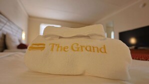 Una toalla doblada con el nombre 'The Grand' bordado, sobre una cama en una habitación de hotel.