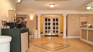 Un vestíbulo de hotel con una mesa de recepción, un área de espera con sillas, suelo de baldosas y una puerta que da a otra habitación.