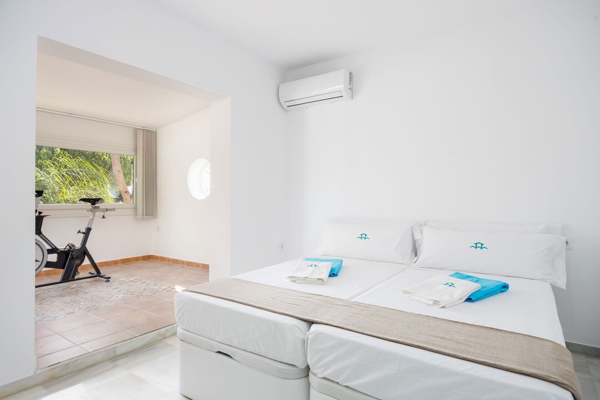 Une chambre à coucher avec un lit, deux oreillers, une fenêtre, un climatiseur et un vélo dans un coin.
