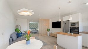 Una cuina moderna amb una illa central, una zona de menjador amb taula rodona, i un espai de sala d'estar amb sofà i plantes.