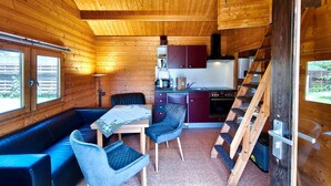 Interno di una baita con soffitto in legno, zona pranzo con tavolo e sedie, cucina con piano cottura e pensili, e una scala in legno.