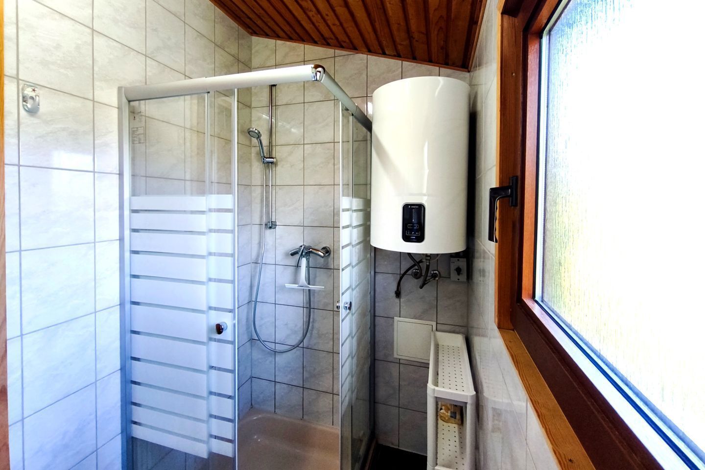 Una ducha con puerta de vidrio, un calentador de agua blanco y una ventana con detalles de madera.
