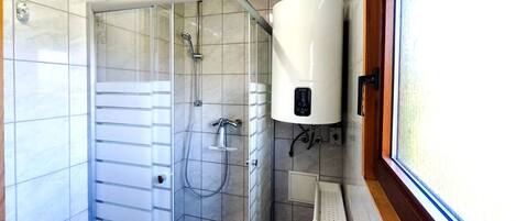 Una ducha con puerta de vidrio, un calentador de agua blanco y una ventana con detalles de madera.