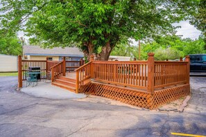 Una terraza de madera con una escalera que lleva a un espacio cubierto, una parrilla y un árbol que da sombra.