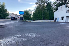 Un parking délimité par une clôture blanche, avec un bâtiment bleu et une voiture noire.