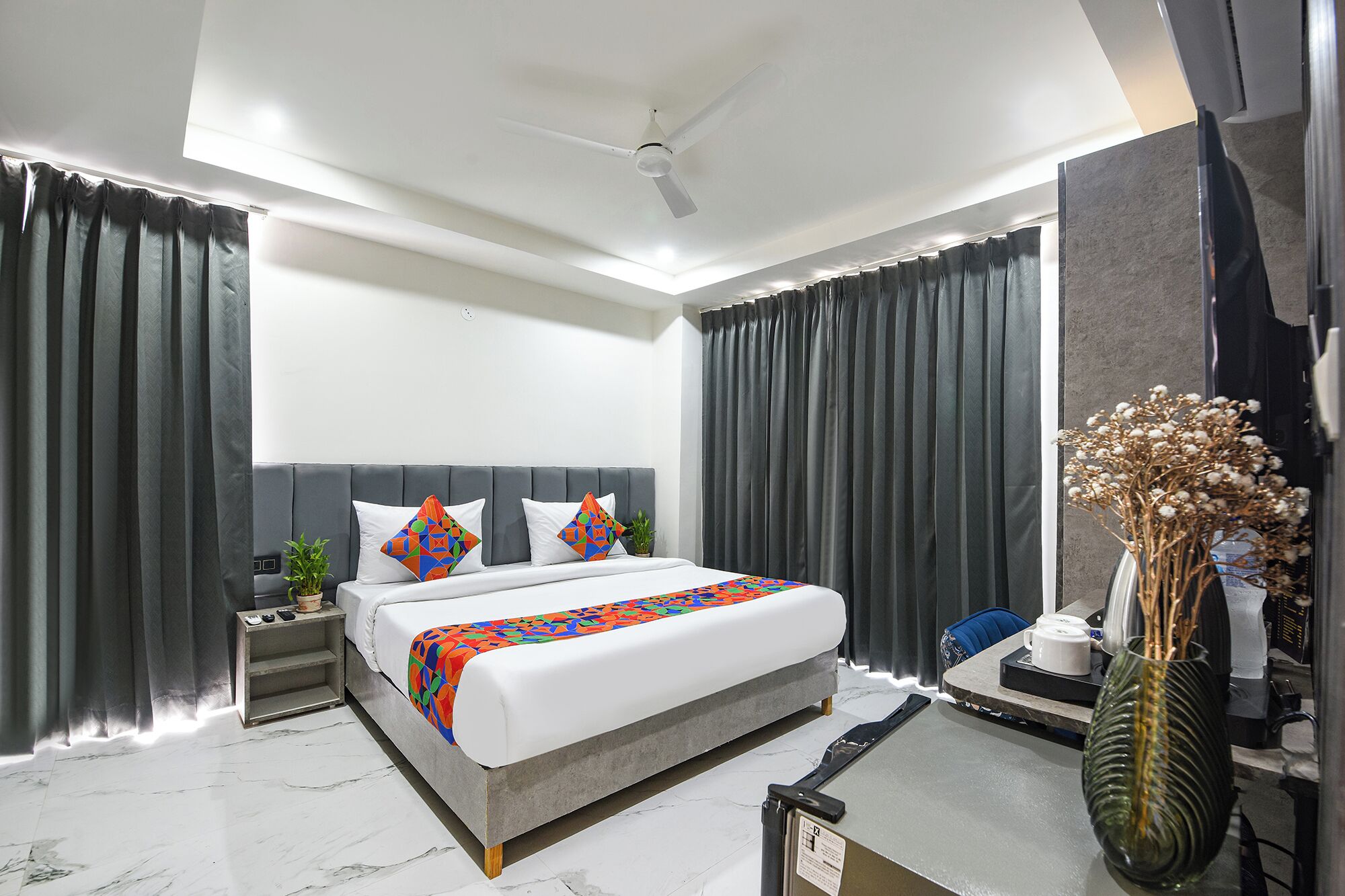 A modern hotel room with a large bed, grey curtains, a ceiling fan, a nightstand with a plant, and a vase with dried flowers.
