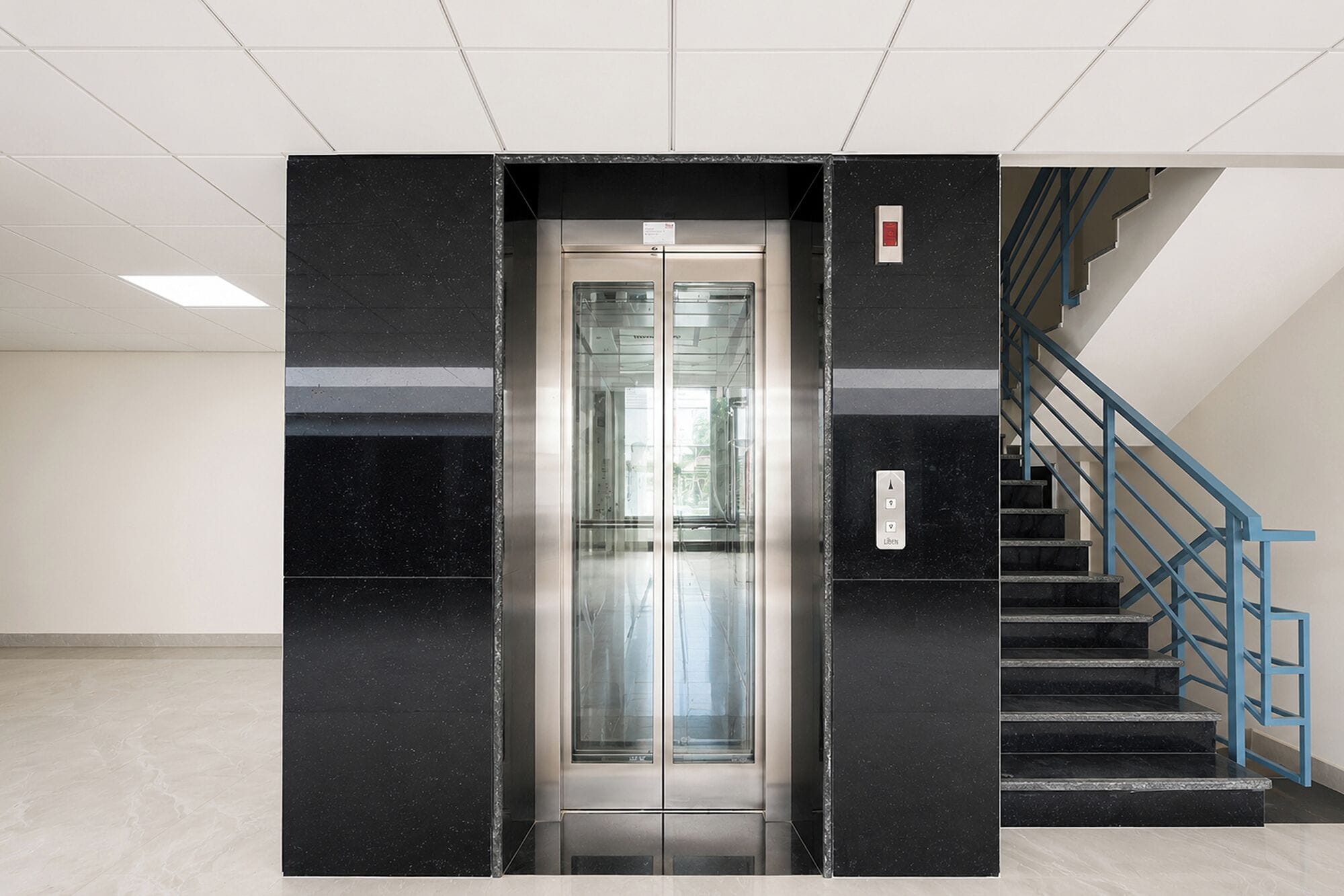 An elevator with a glass door, a staircase with blue railings, and a tiled floor.