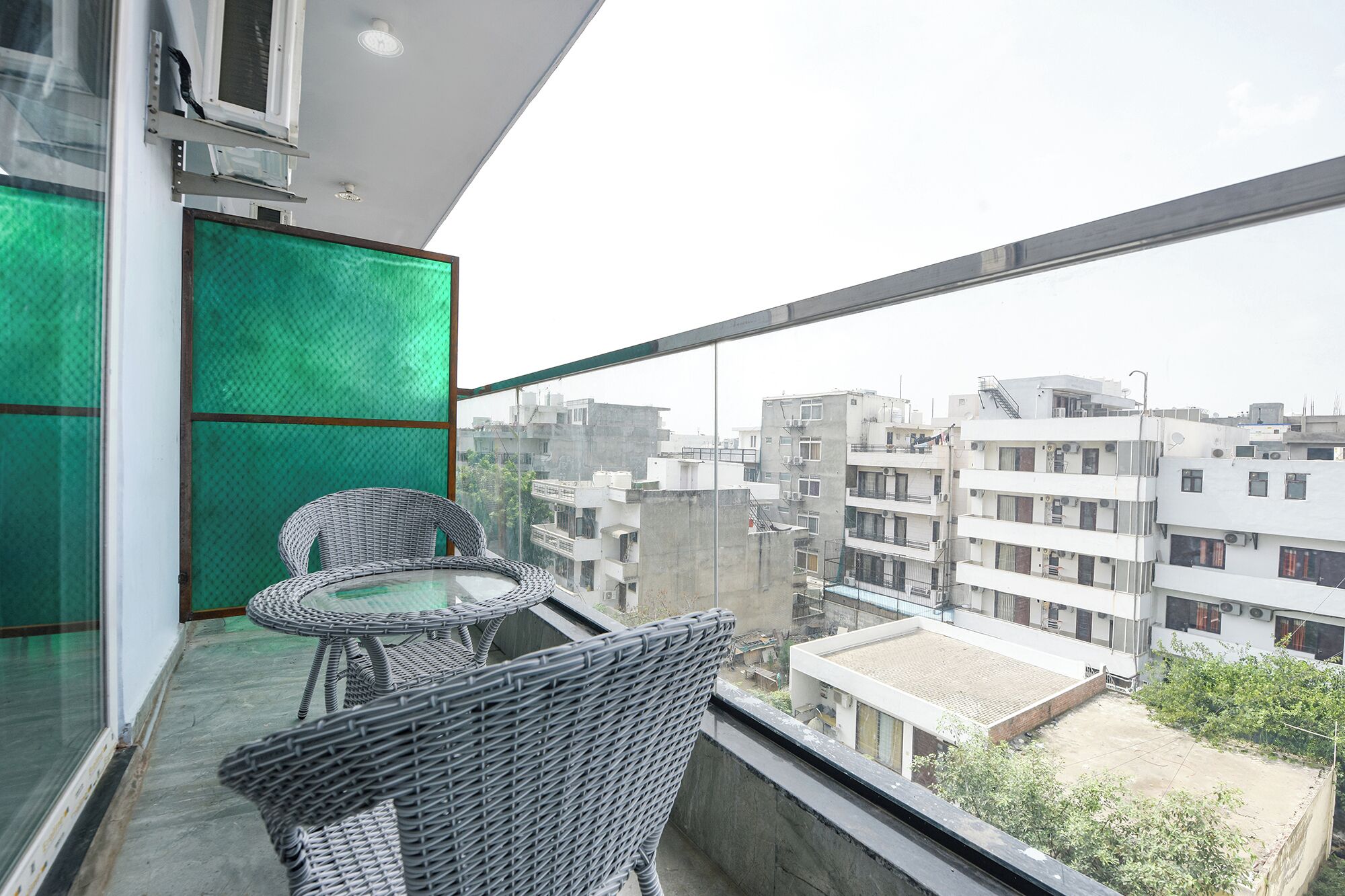 A balcony with wicker furniture and a glass partition, overlooking a cityscape with multiple buildings.