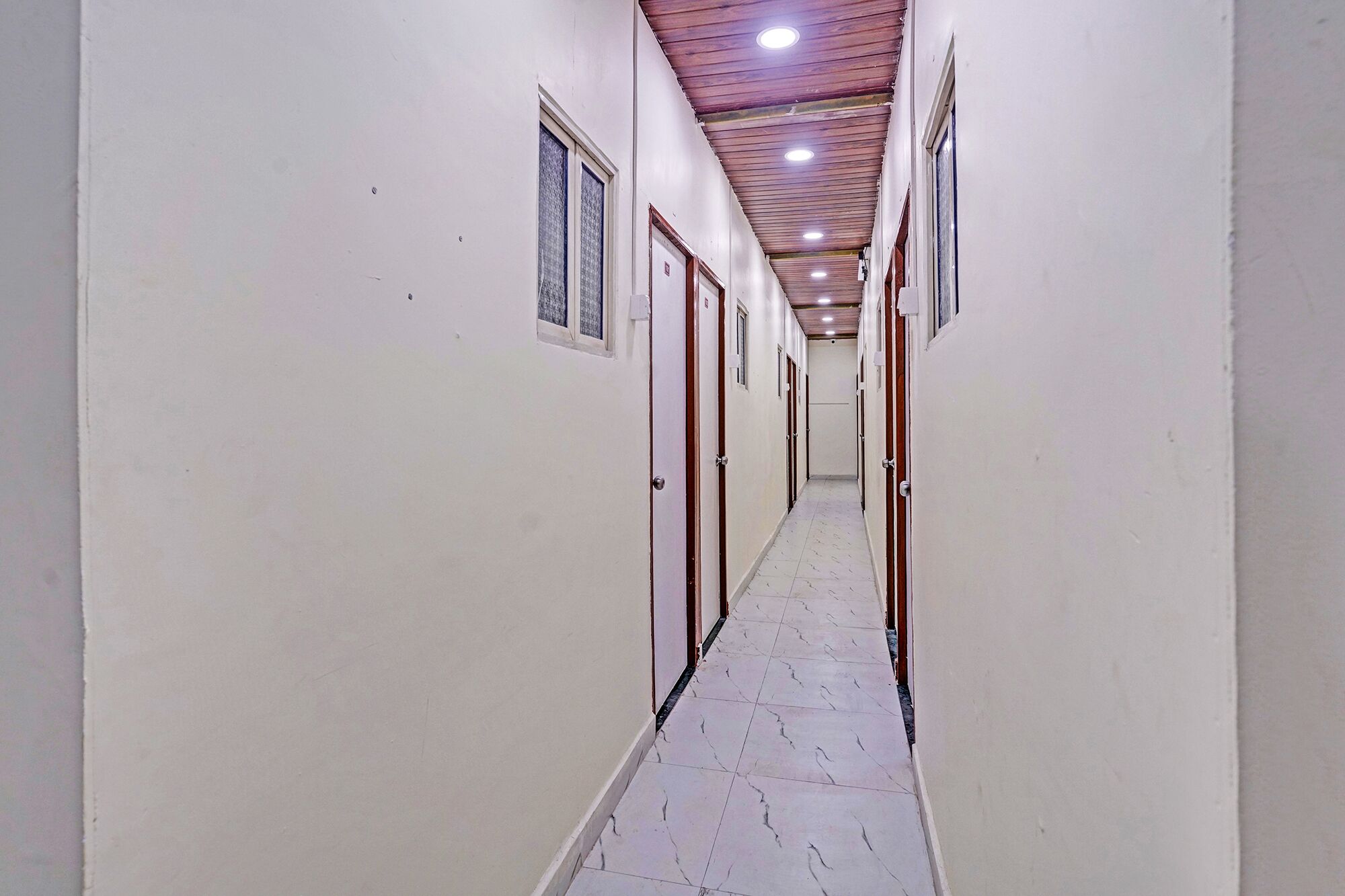 A long, narrow hallway with doors on both sides, a tiled floor, and a wooden ceiling with recessed lighting.
