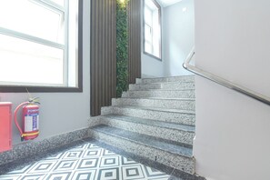 Un escalier orné d’un motif géométrique en carreaux, un extincteur et une fenêtre donnant sur un paysage verdoyant.