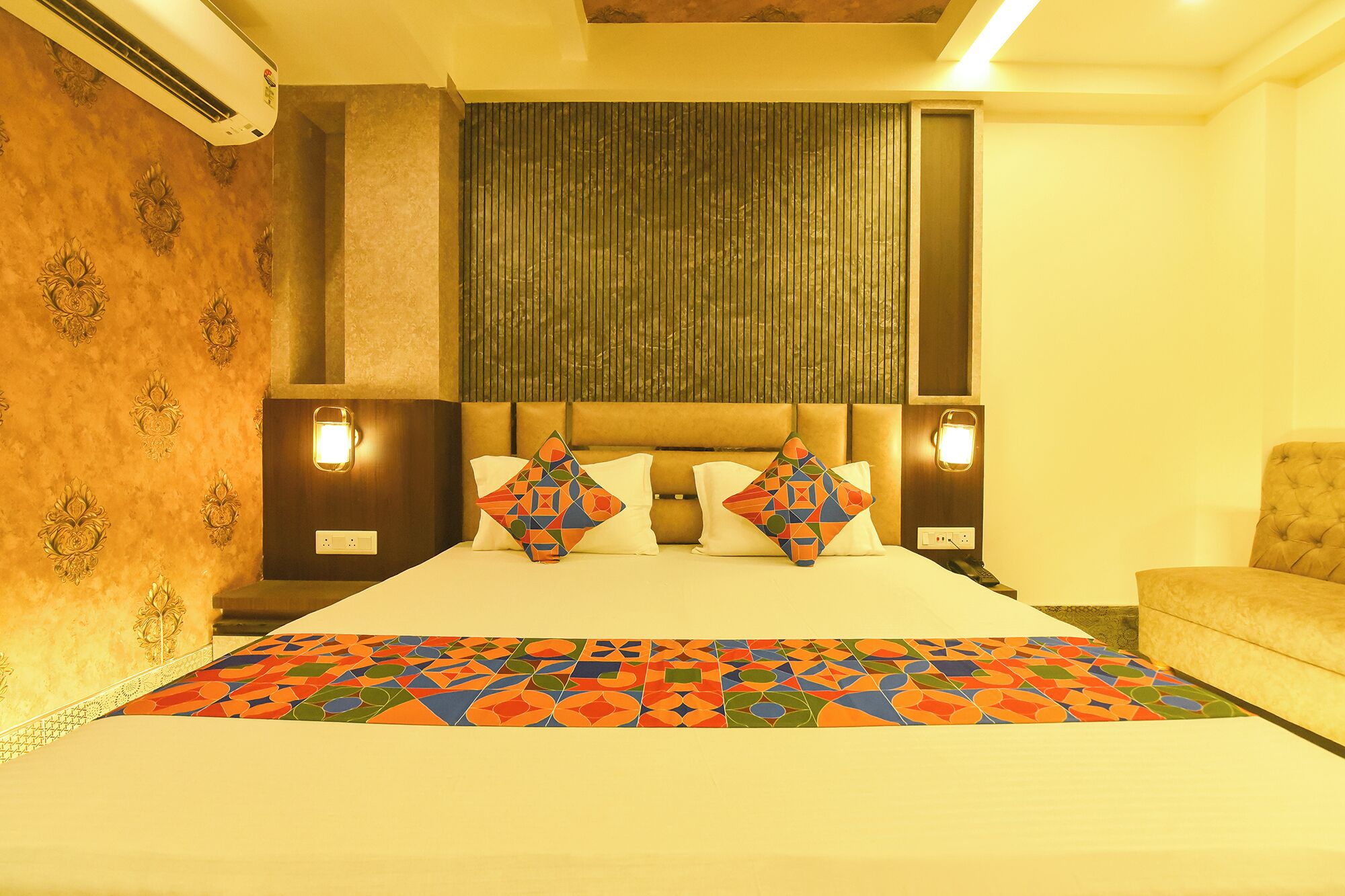 Una habitación de hotel con cama, cabecera, dos lámparas de noche, sofá y colcha estampada.