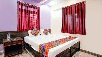Quarto de hotel com uma cama, mesa de cabeceira, garrafa de água e cortinas vermelhas.