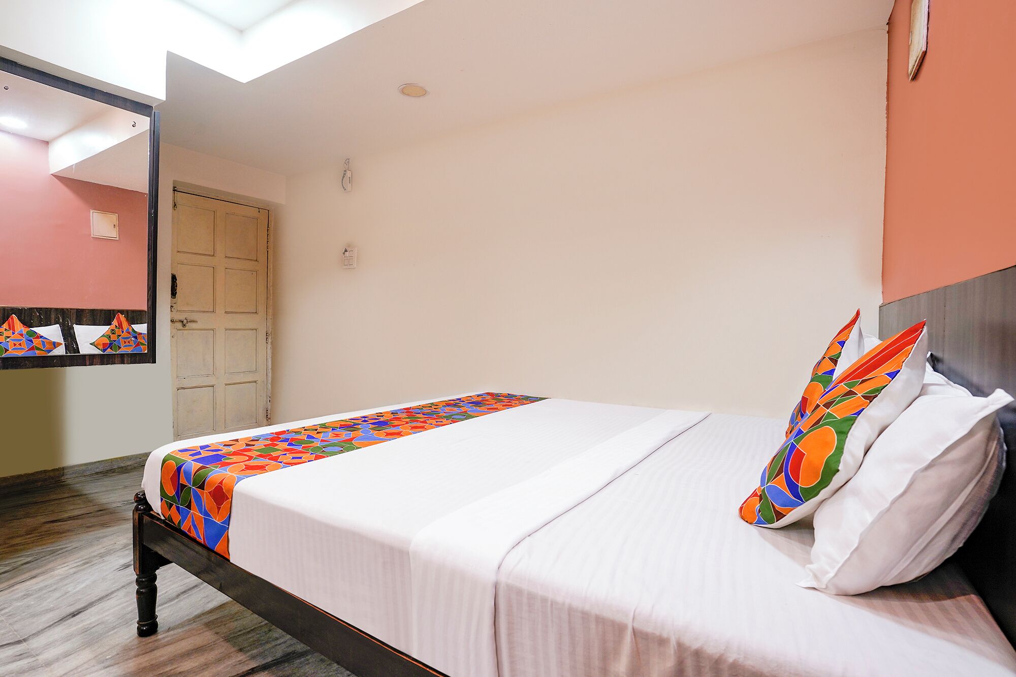 Una cama individual con una colcha de diseño geométrico y colores vivos, un almohadón blanco y otro similar. El cuarto tiene piso de madera, pared blanca y una puerta con un espejo arriba.