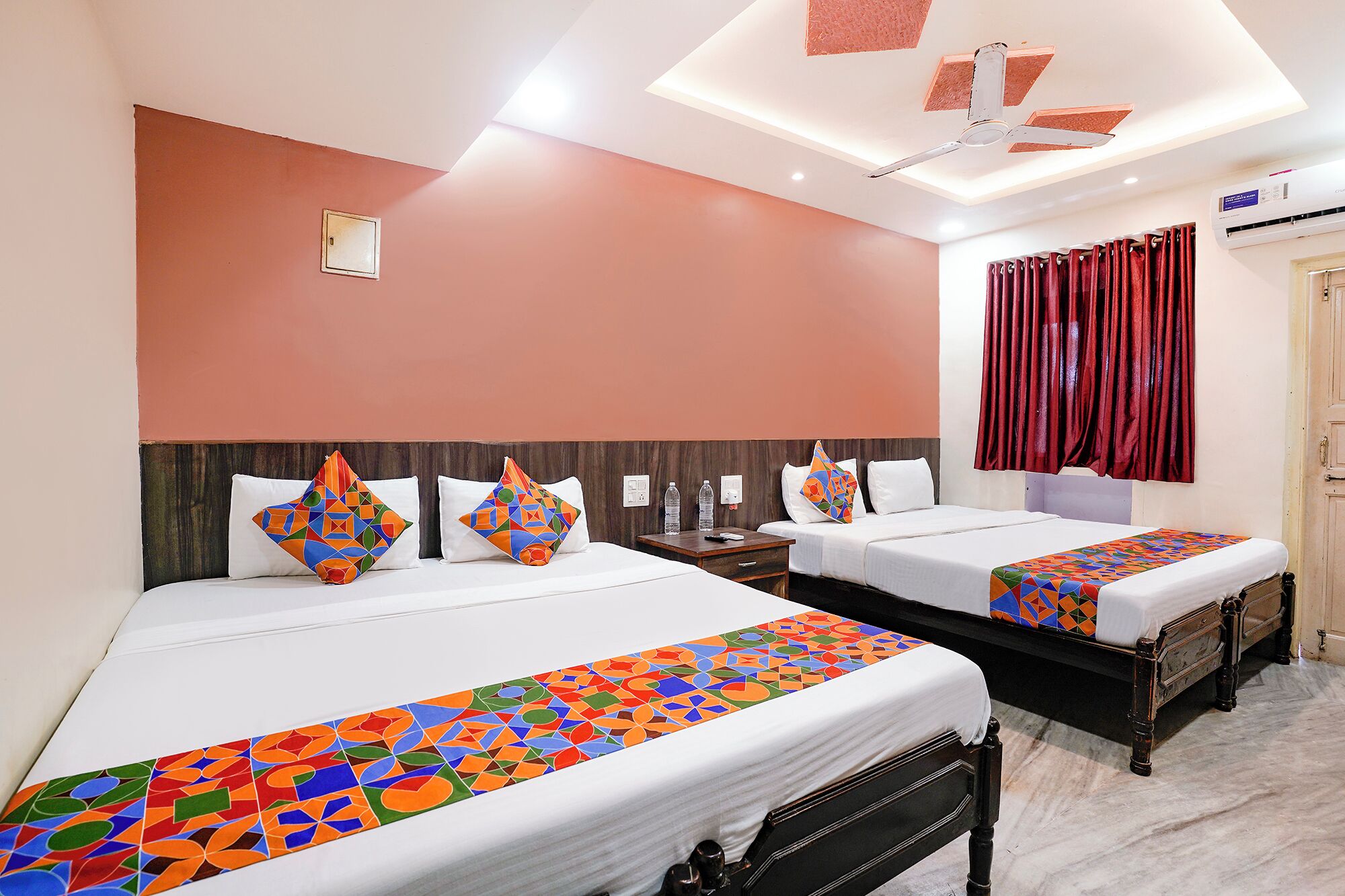 A hotel room with two beds, a ceiling fan, a nightstand, and a window with red curtains.