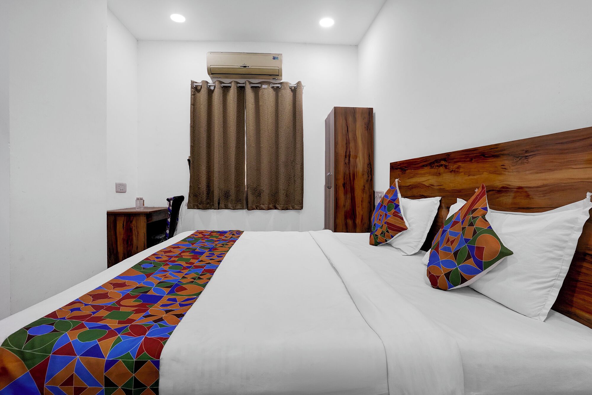 A hotel room with a bed, a wooden headboard, a window with curtains, and an air conditioning unit.
