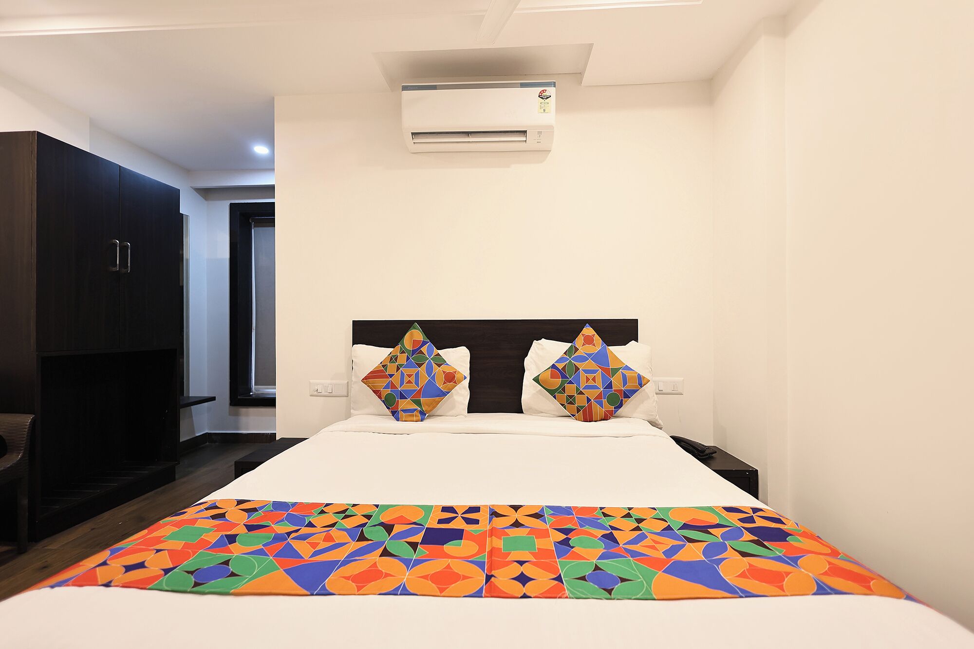 A hotel room with a bed, a nightstand, a wall-mounted air conditioner, and a dark wooden wardrobe.