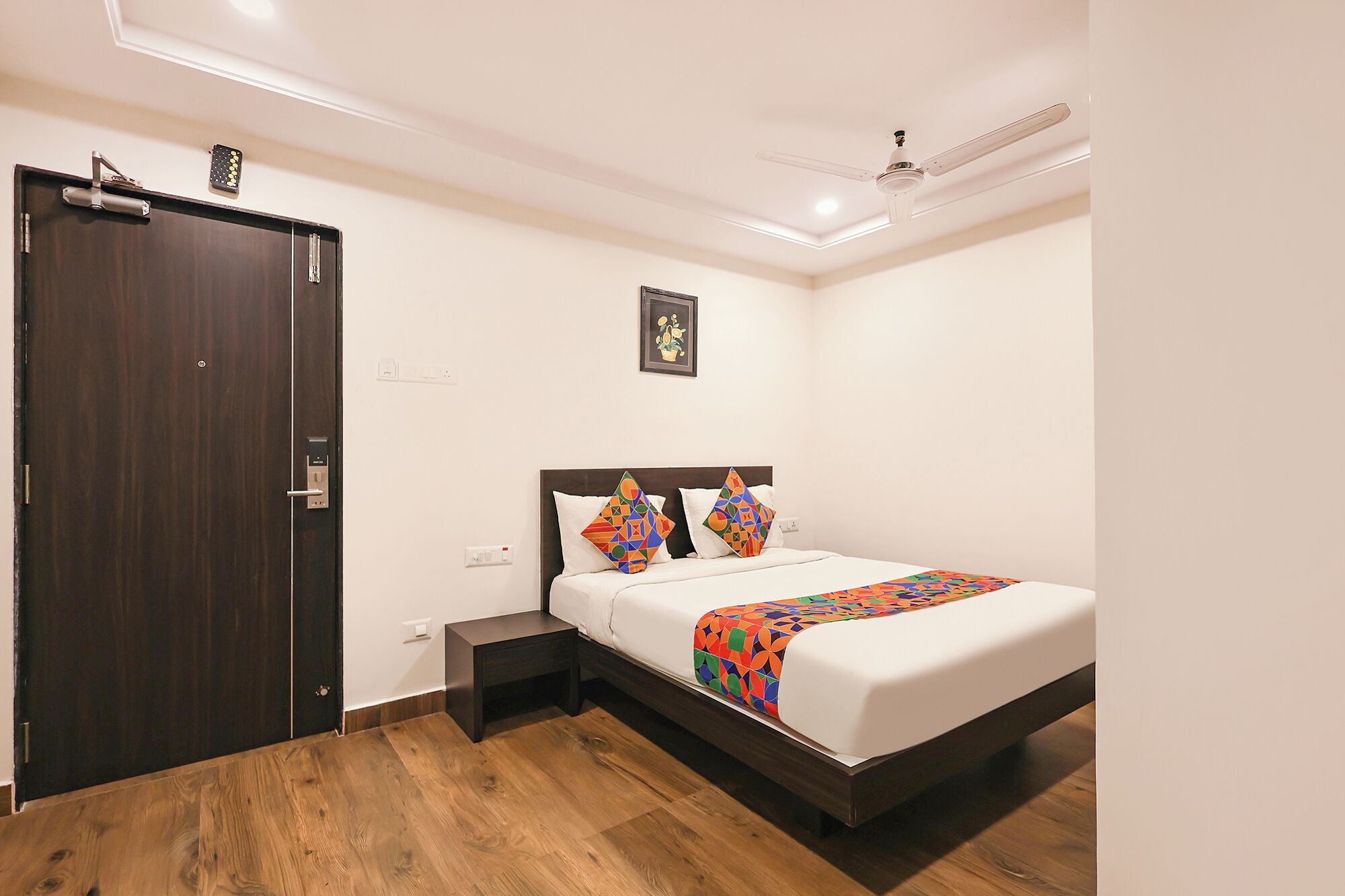 A bedroom with a bed, a nightstand, a wooden floor, a dark door, and a ceiling fan.