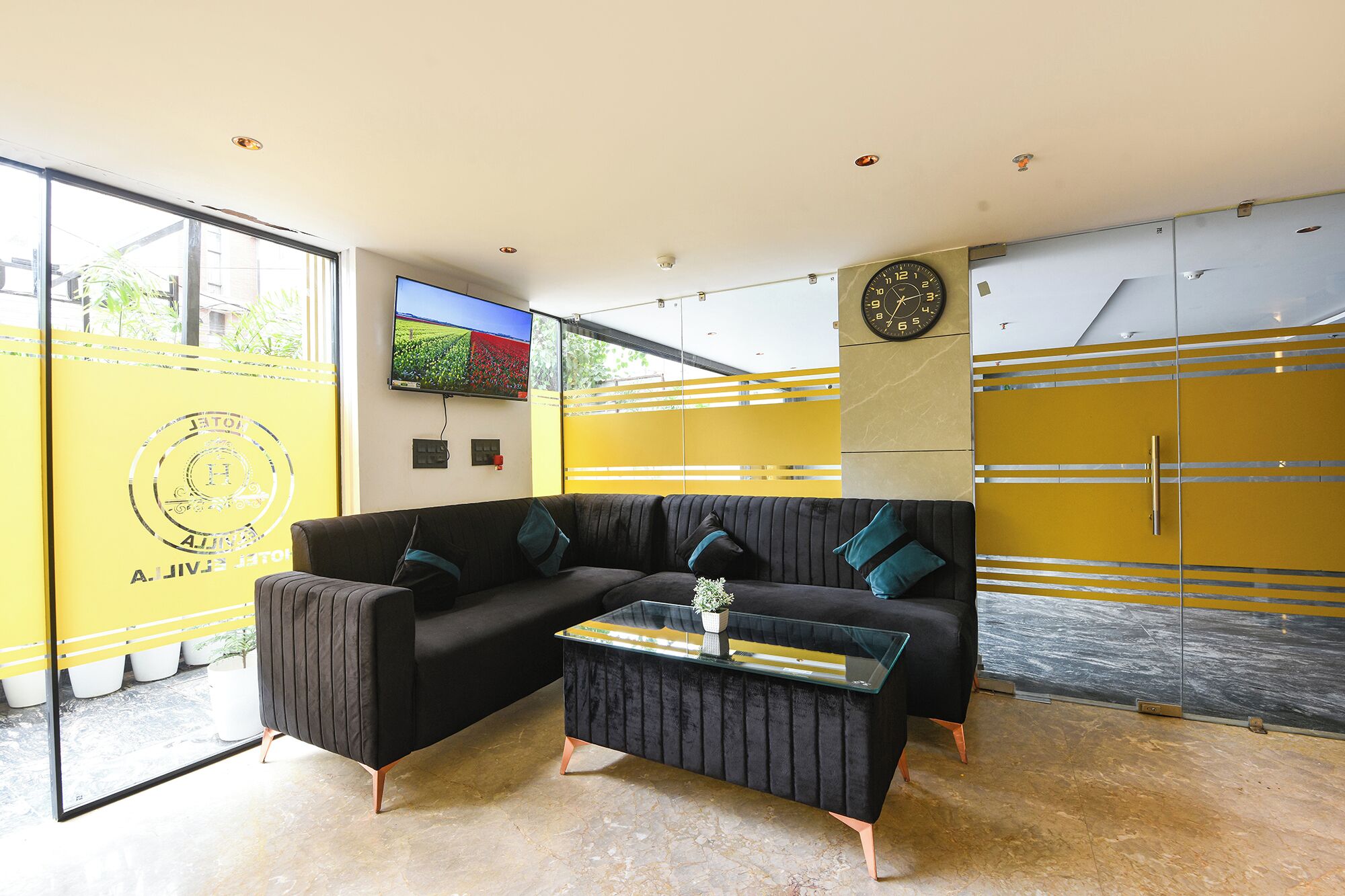A modern living room with a black sofa, a glass coffee table, and a yellow accent wall.