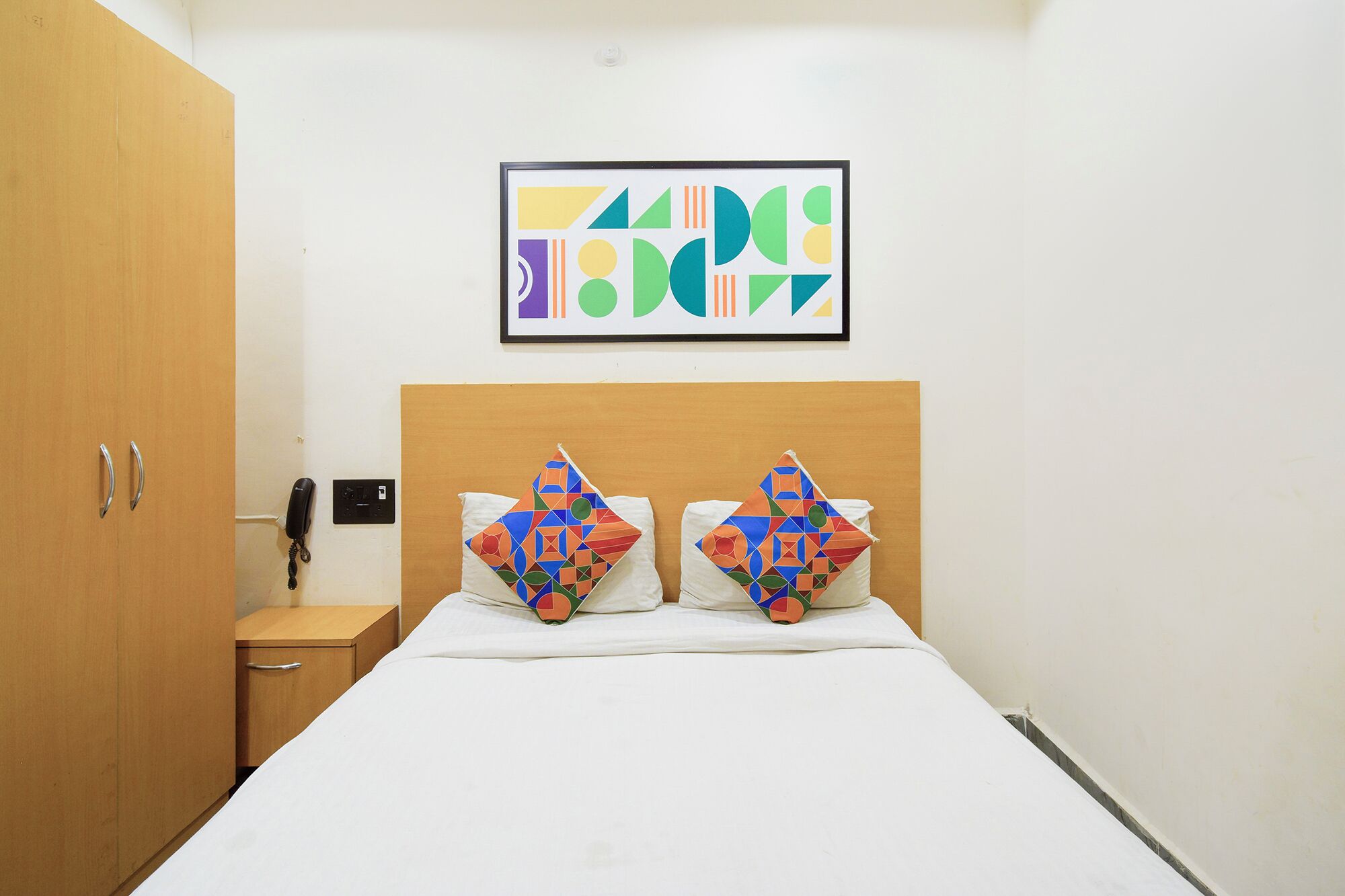 Ein Hotelzimmer mit einem Bett, zwei Kissen mit geometrischen Mustern, einem Holzschrank, einem Nachttisch mit Telefon und einem gerahmten abstrakten Kunstwerk an der Wand.