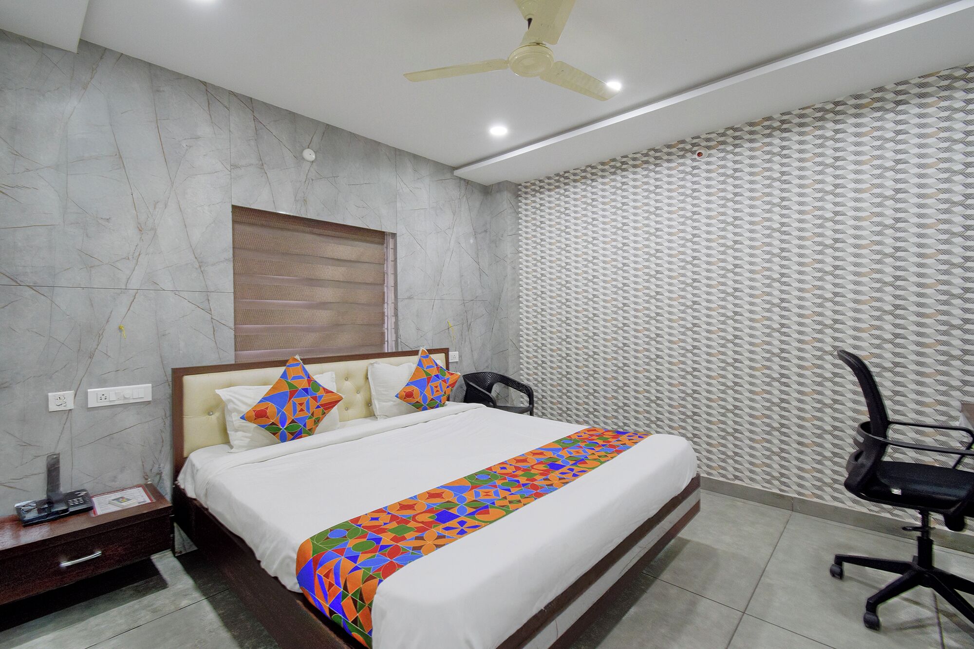 A hotel room with a bed, a desk, a chair, a ceiling fan, and a patterned wall.