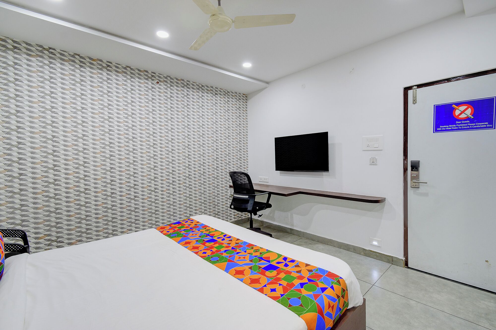 A hotel room with a bed, a desk, a chair, a television, a door, and a ceiling fan.