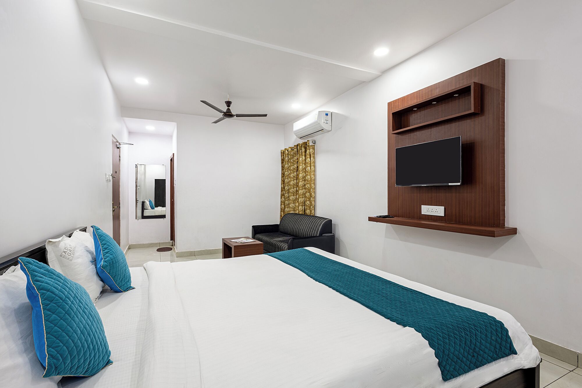 A hotel room with a bed, a TV mounted on the wall, a ceiling fan, and an air conditioning unit.