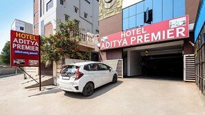 Une voiture blanche est garée devant l’hôtel Aditya Premier, offrant une vue dégagée sur l’entrée et sur un bâtiment dont les murs extérieurs sont peints en bleu et en marron.
