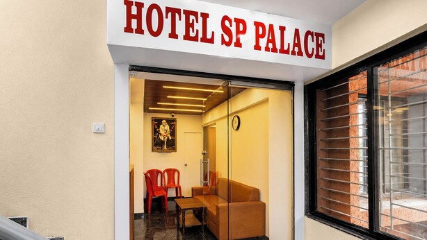 A hotel entrance with a sign reading 'HOTEL SP PALACE,' a glass door, a clock, a red chair, a brown sofa, and a window with blinds. - Fabhotel Sb Palace (Nashik)