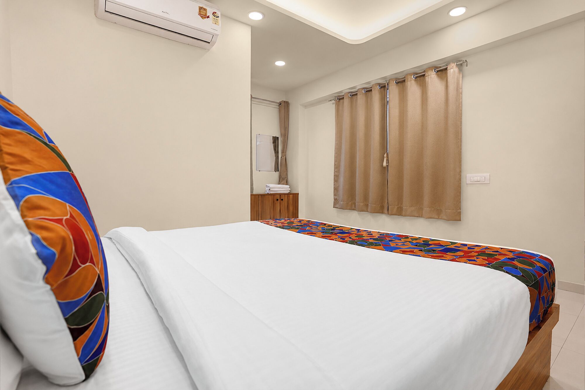 Une chambre d’hôtel avec un lit, une couvre-lit colorée, un oreiller blanc, une table de chevet en bois, un miroir et une fenêtre avec des rideaux.
