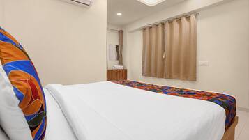 Une chambre d’hôtel avec un lit, une couvre-lit colorée, un oreiller blanc, une table de chevet en bois, un miroir et une fenêtre avec des rideaux.