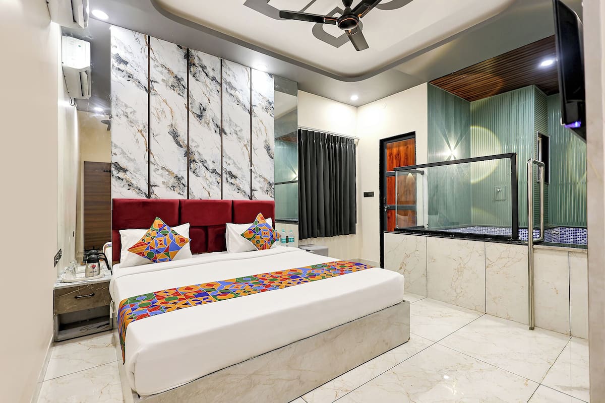 Quarto moderno com parede em estilo marmorizado, cabeceira estofada vermelha, cama grande com almofadas coloridas e banheiro com box de vidro.