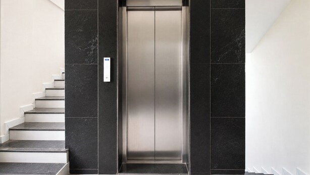 Un elevador moderno con un acabado metálico elegante, flanqueado por paredes de mármol oscuro y escaleras blancas.