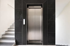 A modern elevator with a sleek metallic finish, flanked by dark marble walls and white staircases. - FabHotel Fortune Inn I (Pune)