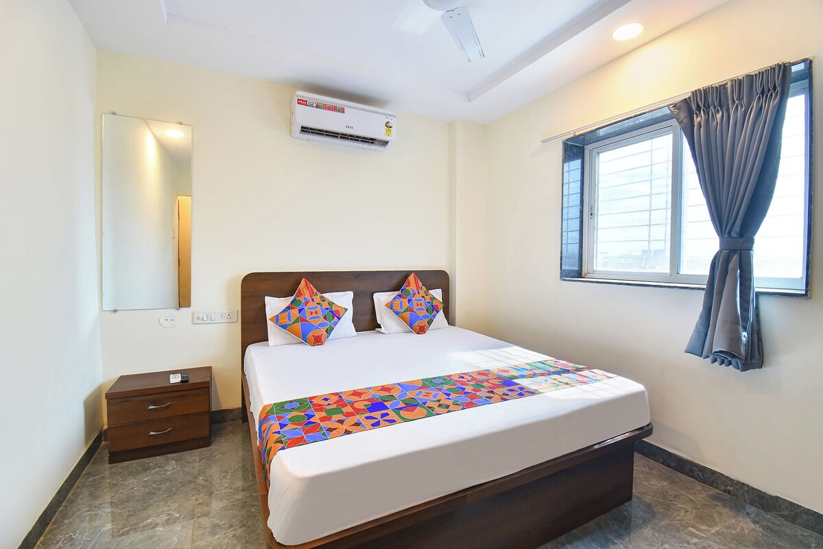 A hotel room with a bed, a nightstand, a mirror, an air conditioning unit, a window with curtains, and a view of the city.