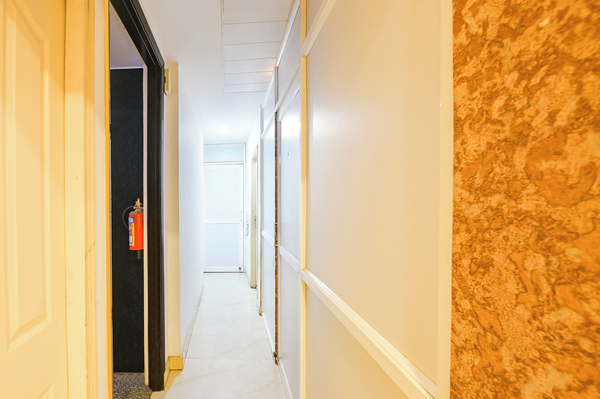 A hallway with white walls, a marble-like textured wall on the right, and a fire extinguisher on the left.