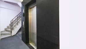 Un ascensor moderno con terminación metálica, una escalera con barandillas de metal y una pared oscura.