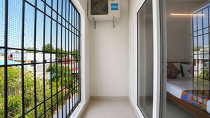 Un pasillo estrecho con una ventana a la izquierda, una cama con almohadas de colores a la derecha y un equipo de aire acondicionado en la pared.
