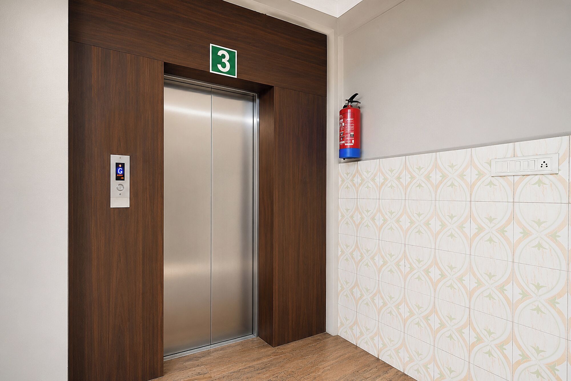 Un ascensor con un letrero verde de salida de emergencia, un extintor montado en la pared y un diseño de baldosas estampadas en la pared contigua.