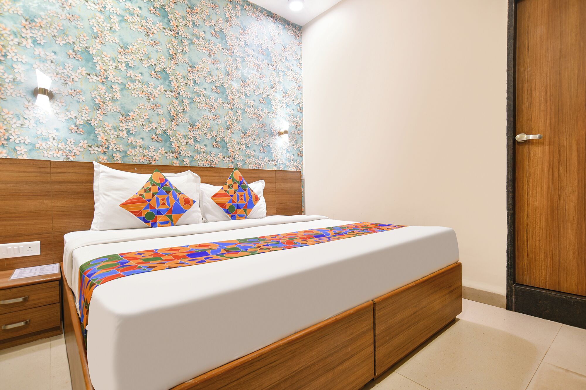 A hotel room with a bed, wooden headboard, white bedding, and a floral-patterned wall.
