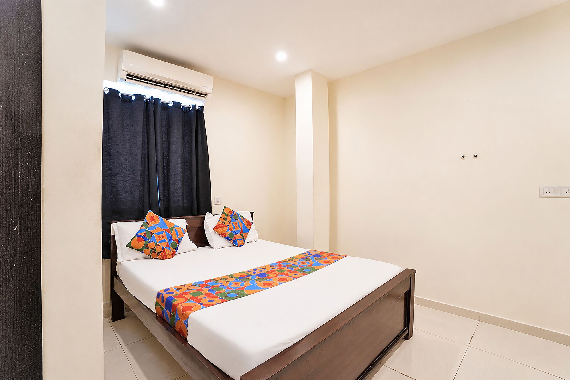 A bedroom with a bed, a window with curtains, an air conditioning unit, and a wall socket.