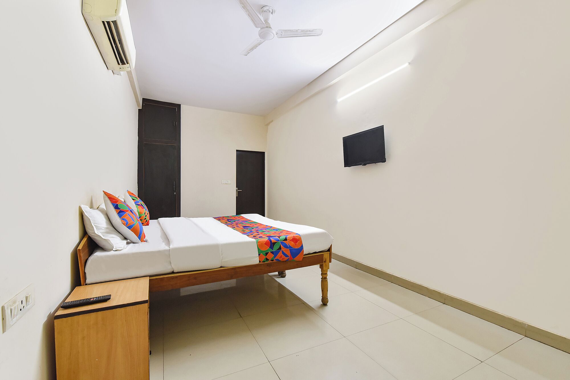 A single bed with a colorful blanket, a wooden nightstand, a ceiling fan, and a TV mounted on the wall.