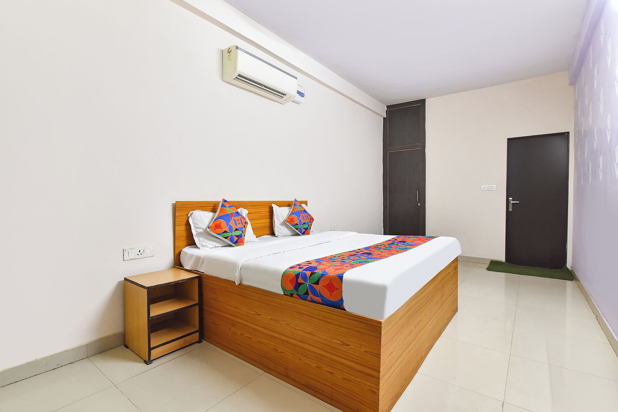 A neatly arranged hotel room with a wooden bed, a small bedside table, a wall-mounted air conditioner, and a door.