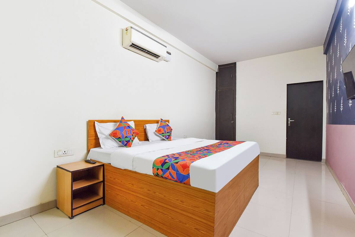 Un dormitorio con cama de madera, ropa de cama blanca, un plaid colorido, una mesita de noche, aire acondicionado instalado en la pared, una puerta oscura y piso de baldosas.