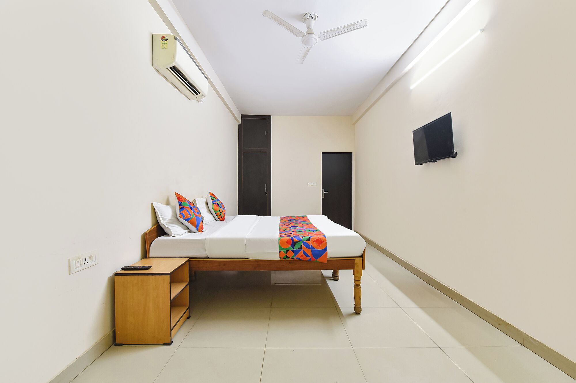 A bedroom with a bed, bedside table, air conditioning unit, ceiling fan, and a TV mounted on the wall.