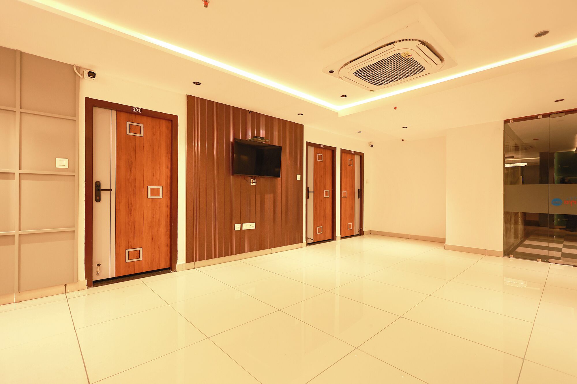 A hotel hallway with wooden doors, a flat-screen TV mounted on the wall, and a tiled floor.