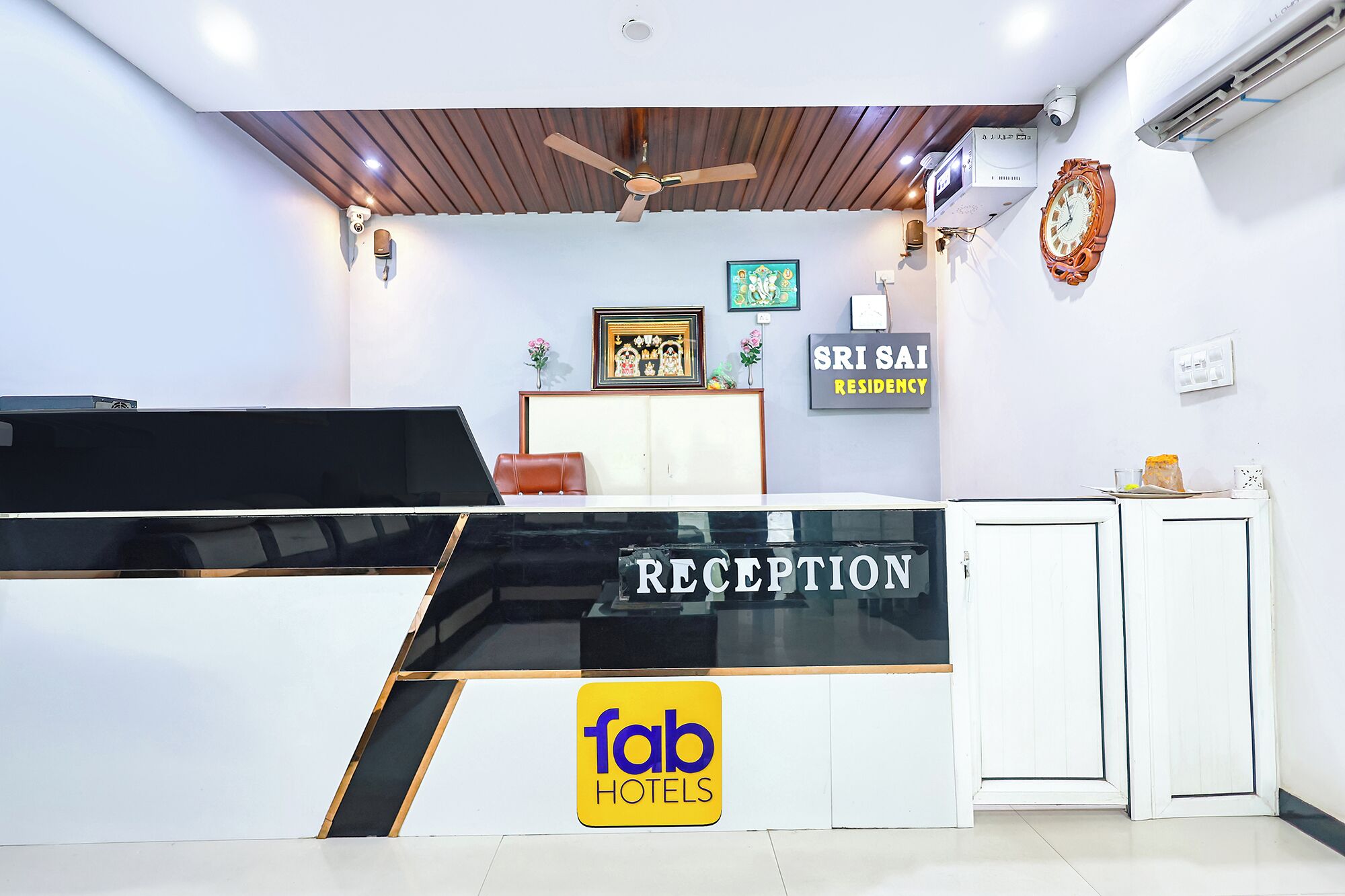 Een receptiebalie met een bordje 'fab hotels', een ingelijst beeld en een bordje 'SRI SAI RESIDENCY'.