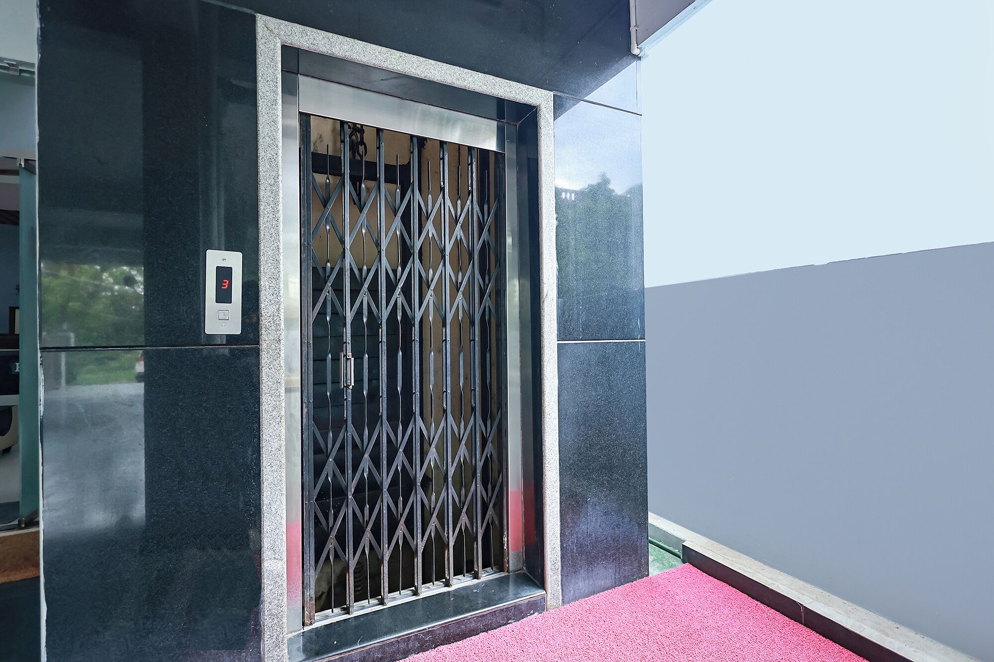 A door with a metal security gate, a digital lock, and a red carpet leading to a building with a grey exterior.