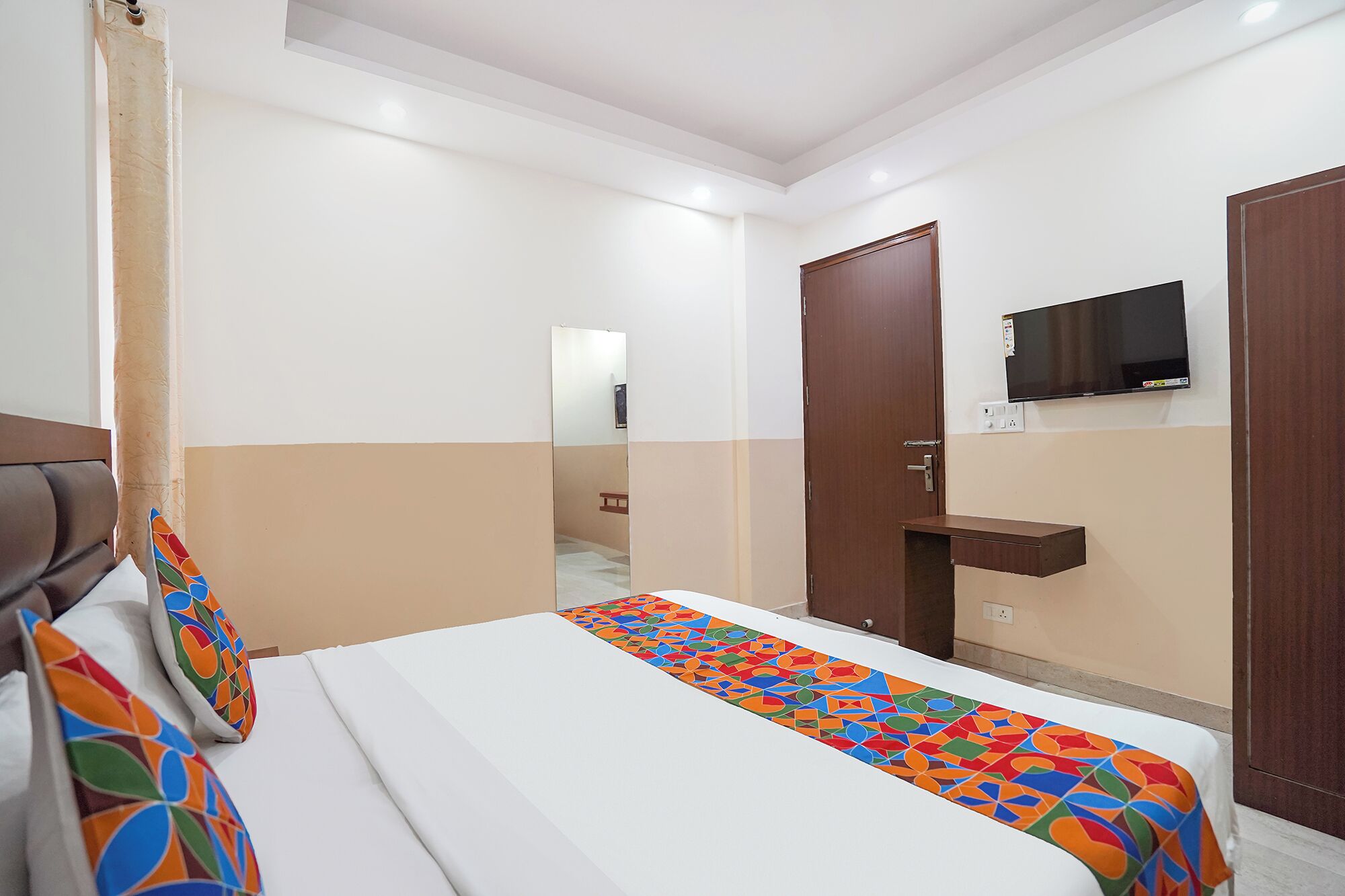 A hotel room with a bed, a bedside table, a mirror, a TV mounted on the wall, and a door.