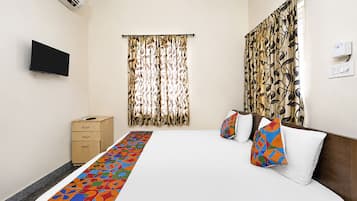 A hotel room with a bed, a TV mounted on the wall, a wooden nightstand, and a window with patterned curtains.